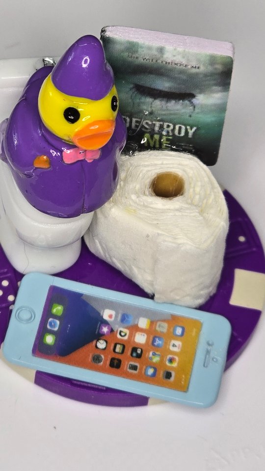 Purple poker chip duck sitting on a tiny toilet next to a miniature phone and book